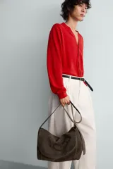 Bolso tipo bandolera de piel con acabado serraje, color marrón oscuro. Presenta un compartimento principal con cierre de solapa y cremallera, y un bolsillo interior con cierre de cremallera. Incluye un asa de hombro ajustable de piel.