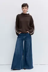 Jersey de punto color marrón chocolate, de cuello alto y corte holgado. Presenta un diseño acanalado y un detalle de costura decorativa en color blanco que recorre la parte superior de las mangas.