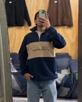 Buzo de polar térmico con diseño de bloques de color en azul marino y beige. Presenta cuello alto con medio cierre frontal y un bordado decorativo en el pecho. Es una prenda de abrigo ligera y versátil.