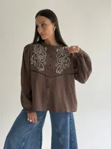 Camisa color marrón chocolate con bordado artesanal de flores y hojas en hilo crudo. Presenta cuello con volado, mangas largas con puños abotonados y cierre frontal con botones.