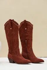 Bota texana de gamuza color marrón, con taco y puntera fina. Presenta bordados decorativos en el cuerpo de la caña y detalles de tachas metálicas en la parte superior y en la costura inferior.