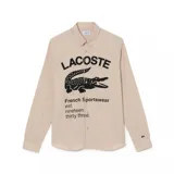 Camisa de manga larga Lacoste en color beige, con cuello abotonado y cierre frontal de botones. Presenta un estampado gráfico grande en el frente con el nombre de la marca, el logo del cocodrilo y la leyenda 'French Sportswear est. nineteen thirty three'. Incluye un pequeño logo bordado en el puño de la manga izquierda.