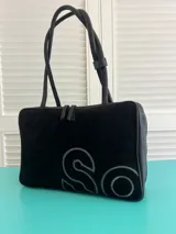 Bolso de mano o al hombro, tipo tote bag, color negro. El cuerpo principal es de gamuza con un gran logo estampado en contorno gris claro. Las asas y los bordes son de cuero liso negro.