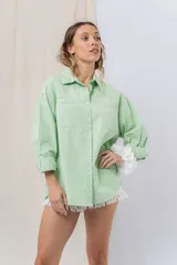 Camisa verde pastel de algodón, corte oversize, con cuello camisero, cierre con botones, mangas largas con puños con volados de encaje blanco, dos bolsillos frontales con solapa y ruedo irregular. 