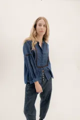 Camisa de jean azul oscuro con cuello mao, botones frontales, mangas largas abullonadas con puños elásticos y detalles bordados en color marrón en los hombros y mangas.