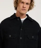 Campera de hombre de jean negro, modelo regular, con cuello inglés, abotonado frontal, dos bolsillos con solapa en el pecho y manga larga ajustada.