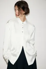 Camisa de satén color blanco, de corte clásico con cuello solapa, manga larga con puños y cierre frontal con botones dorados.