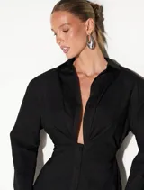 Vestido camisero corto negro, de manga larga con puños anchos, escote en V profundo y detalle de frunces en el busto. Cuenta con cierre frontal de botones y silueta entallada.