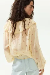 Blusa de gasa arrugada de corte holgado, en tono amarillo claro con estampado floral. Cuenta con escote en V profundo, un amplio volante ribeteado con encaje alrededor del cuello y cordones finos para atar al frente. Mangas largas con puños elásticos.