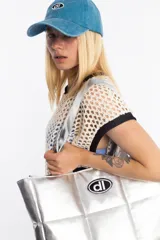 Bolso shopper plateado acolchado, con diseño puffer y costuras a cuadros. Cuenta con tiras para el hombro y un parche ovalado negro y blanco con las iniciales dl en el centro.