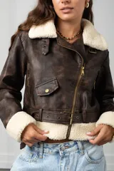 Chaqueta de estilo biker confeccionada en eco cuero con efecto gastado, presenta cuello y puños con forro de corderito sintético. Incluye cierre frontal cruzado, bolsillo pequeño con solapa y cinturón ajustable en la cintura.