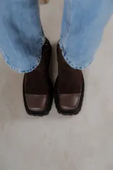 Bota corta de gamuza marrón con puntera de cuero liso, suela track negra y correa desmontable con tachas metálicas.