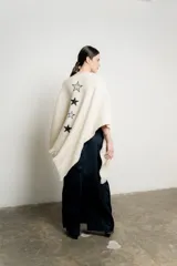 Ruana de tejido de punto color crudo con terminaciones de flecos en los bordes. Presenta apliques de estrellas bordadas a mano en diferentes tamaños y tonos.