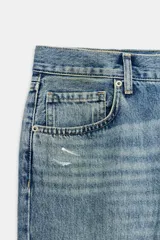 Jeans de tiro medio con corte recto y largo completo. Presentan un diseño de cinco bolsillos, cierre frontal con cremallera y botón, y un acabado con efecto lavado. Incluyen detalles de roturas y parches decorativos en la parte delantera.