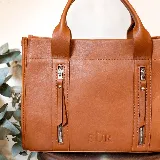 Cartera de cuero color tostado, estructurada, con doble asa de mano, correa desmontable y dos bolsillos frontales verticales con cierre. Presenta detalles de costuras pespunteadas y el logo de la marca grabado en la parte inferior.
