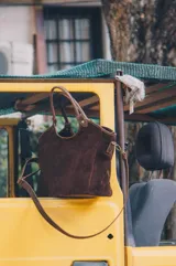 Cartera tipo tote bag de gamuza color marrón, con doble asa de mano y correa larga ajustable y desmontable. Cuenta con herrajes metálicos en tono bronce.