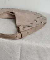 Cartera tipo hobo de símil gamuza color beige, con diseño de media luna y decorada con tachas metálicas plateadas en todo su contorno. Incluye correa ajustable y bolsillo interno.