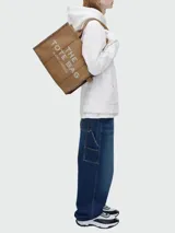 Bolso tipo tote de gran tamaño en color beige, confeccionado en lona resistente. Presenta el logo estampado en negro en el frente y detalles de cintas laterales con inscripciones. Cuenta con doble asa superior para llevar al hombro.