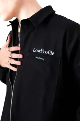 Camisa de gabardina negra de diseño unisex y corte holgado. Cuenta con un cierre frontal de doble camino, cuello camisero y detalles de estampa y bordado en el pecho con el texto 'Low Profile'.