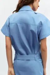 Vestido corto de estilo camisero color azul, con cuello solapa, manga corta y cierre frontal con botones. Presenta un bolsillo de plastrón con solapa en el pecho y un cinturón ajustable del mismo tejido en la cintura.
