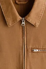 Campera de algodón color marrón con cuello camisero, cierre frontal, bolsillo superior con cierre y bolsillos laterales ribeteados.