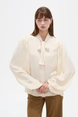 Camisa de poliéster color marfil, de diseño holgado, con cuello alto lazada y mangas largas abullonadas.