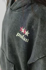 Canguro gris oscuro con efecto lavado (acid wash) y capucha. Presenta un pequeño estampado frontal con estrellas y la palabra "pulau" en tonos rosado y amarillo. Tiene bolsillo canguro y puños acanalados.