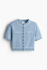 Cárdigan corto de punto azul celeste, manga corta, con cuello redondo y cierre frontal de botones metálicos. Presenta ribetes blancos contrastantes en el cuello, puños, bolsillos superiores decorativos y ruedo inferior.