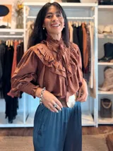Blusa blanca de estilo romántico con volados tipo shabot en el frente, cuello alto con lazo ajustable y mangas largas abullonadas con puños ajustados.