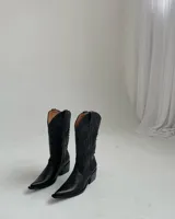 Bota estilo texana de caña alta, confeccionada en cuero negro con forro interior de cuero. Presenta bordados decorativos en la caña y en la puntera, con taco cuadrado de altura media y diseño sin cierre.