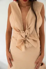 Vestido largo color beige, de silueta ajustada, con escote en V profundo, sin mangas, hombros marcados y lazo en la cintura. La falda recta cuenta con terminación de flecos en el ruedo.