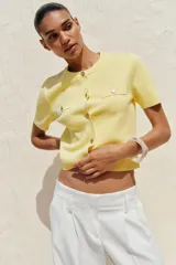 Chaqueta de punto color amarillo pálido, con cuello redondo, manga corta, cierre frontal con botones dorados y bolsillos de parche en el pecho.