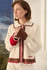 Cardigan de punto color marfil, con cuello tejido a mano en crochet marrón, cierre frontal de botones y detalles en marrón y rojo en los puños y el ruedo. Presenta mangas semi oxford y bordado de la marca en el bajo.