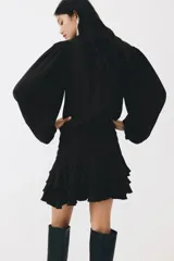 Vestido corto negro de mezcla de viscosa, con escote redondo, abertura frontal con tiras finas y mangas largas abullonadas con puños elásticos. Presenta sección entallada en la cadera con alforzas y falda corta con volados escalonados.