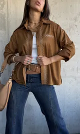 Camisa de gamuza color camel, con cuello clásico, cierre frontal de botones y detalle de bordado geométrico en color crema a lo largo de la botonadura y en el bolsillo frontal. Presenta mangas largas con puños doblados.