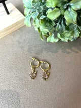 Aros tipo argolla pequeña en tono dorado con dije colgante en forma de racimo de uvas, decorado con pequeñas piedras brillantes en tonos violeta y morado.