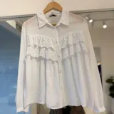 Camisa blanca de manga larga con cuello camisero, cierre frontal con botones y detalles de volados con encaje broderie en el pecho.