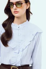Camisa celeste de manga larga con sutiles rayas verticales, cuello alto mao fruncido y cierre frontal con botones. Presenta volados decorativos en los hombros y el frente, y puños elásticos fruncidos.