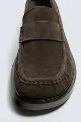 Mocasín de vestir para hombre, fabricado en piel con acabado serraje (gamuza) color marrón oscuro/café. Presenta un detalle de antifaz en el empeine con costuras visibles y horma cuadrada. La suela es negra y a contraste con un ligero tacón.