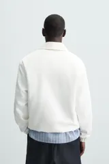 Cazadora tipo canguro de calce relajado, color blanco roto o crema, confeccionada en mezcla de lyocell y algodón. Presenta cuello solapa con cierre frontal de botonadura oculta, manga larga con puños elásticos y bolsillos frontales de vivo. El bajo es ajustable con elástico oculto en los laterales.