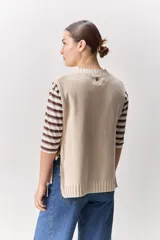 Chaleco tejido de punto color beige, con cuello redondo, terminaciones en rib y lazos laterales regulables que permiten ajustar la prenda.