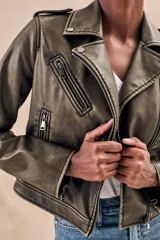 Campera corta color verde oliva de cuero sintético con cuello clásico, cierre frontal asimétrico con cremallera, bolsillos con cremallera y solapa, y detalles de costuras en los hombros.