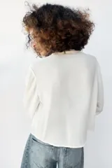Blusa blanca de manga larga con cuello en V, cierre frontal con botones y detalles de bordados florales y encaje en el frente.