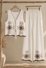 Conjunto beige de dos piezas confeccionado en tela liviana, con detalles bordados en marrón oscuro en los bordes y diseño floral. Incluye un chaleco corto sin mangas con escote en V y tiras para atar al frente, y un pantalón largo de corte recto y holgado.