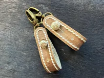 Llavero artesanal de cuero crudo en color tostado, con costuras decorativas blancas, botón de tiento y gancho metálico de bronce envejecido.