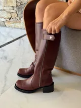 Bota de caña alta estilo biker, confeccionada en cuero color chocolate. Presenta un cierre metálico lateral que recorre toda la caña, estribo y cadena decorativa desmontables, y taco cuadrado con plataforma.