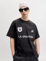 Remera negra de manga corta y cuello redondo, de corte holgado. Cuenta con estampado frontal de estilo deportivo que incluye la inscripción "LA GRACIOSA", un parche circular con diseño de sol y el logo de la marca.
