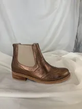 Botín tipo Chelsea de cuero metalizado en tono bronce, con paneles elásticos laterales en color beige. Presenta detalles de costuras decorativas tipo brogue en la puntera y laterales, con suela de goma antideslizante y taco bajo foliado.