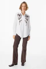Camisa blanca de corte clásico con cuello camisero y cierre frontal de botones. Presenta un bordado lineal en color marrón oscuro en ambos lados del frente, representando motivos florales.