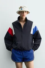 Chaqueta técnica de corte relajado con cuello subido y cierre frontal de cremallera bidireccional. Presenta un diseño color block en tonos azul marino, blanco, rojo y azul eléctrico. Incluye bolsillos frontales de vivo, puños y ruedo elásticos, y forro interior de red.
